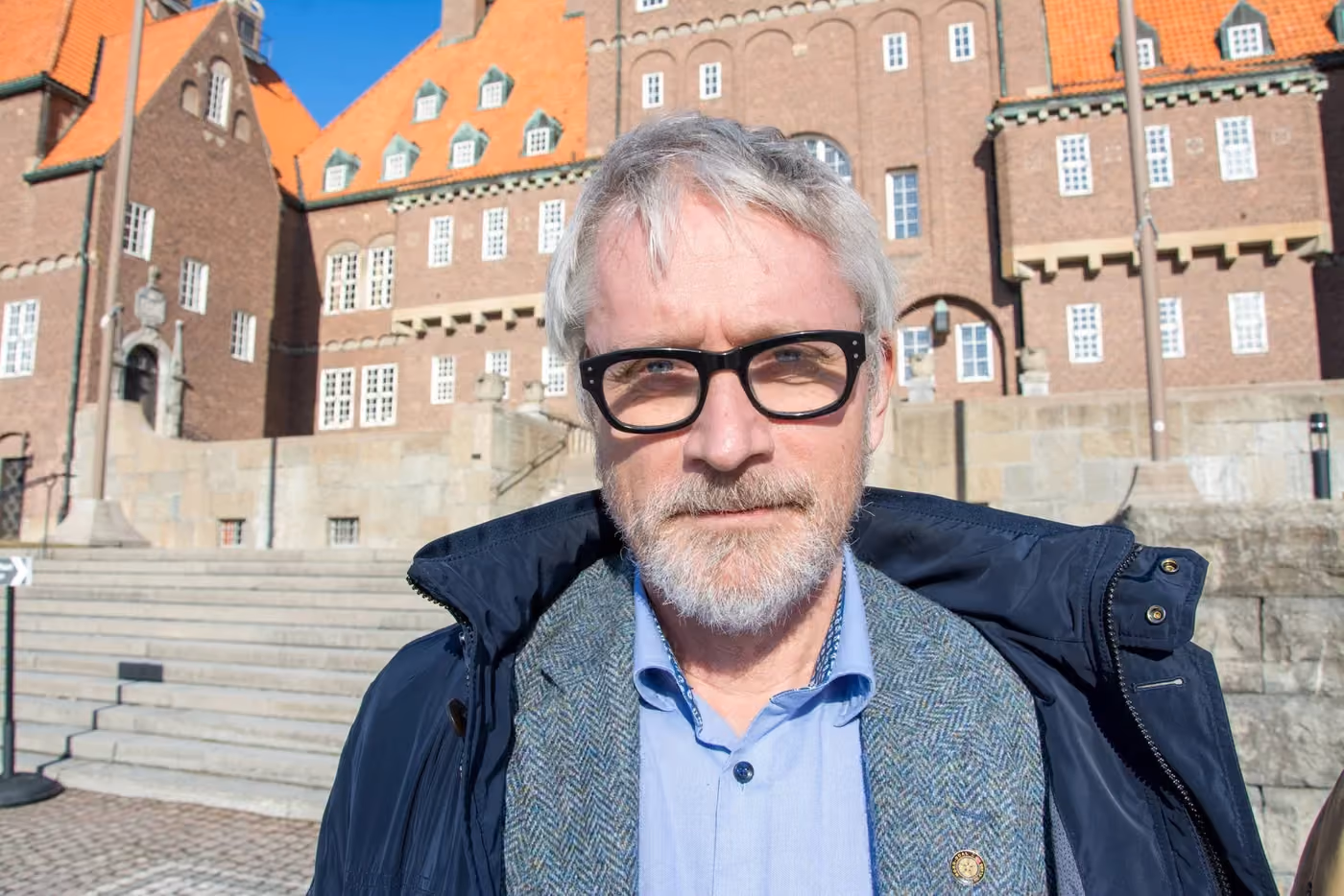 Mann med grått hår, skjegg og briller står foran en bygning med rødt tak i sollys.